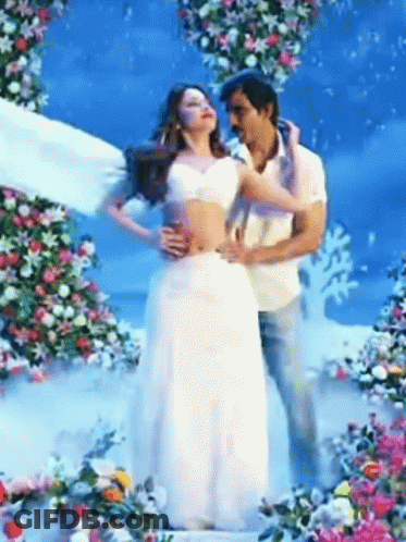 A Man Is Holding A Woman In A White Dress In Front Of Flowers GIF