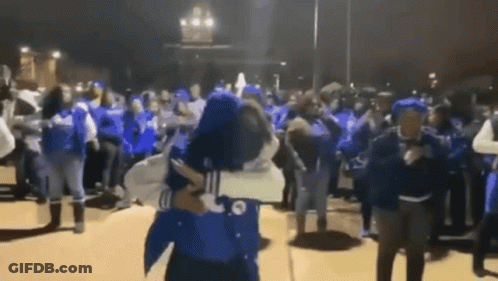 A Man Is Holding A Woman In His Arms In Front Of A Crowd Of People Wearing Blue Shirts . GIF