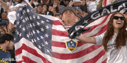 A Man Is Holding An American Flag And A Woman Is Wearing A Shirt That Says Herbalife GIF