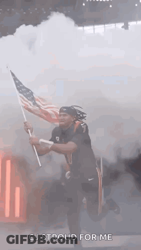 A Man Is Holding An American Flag In Front Of Smoke Coming Out Of A Stadium . GIF