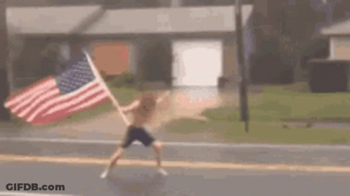 A Man Is Holding An American Flag While Walking Down A Street . GIF