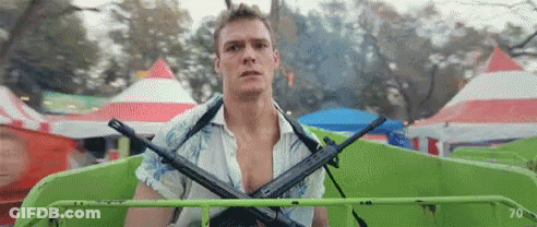 A Man Is Holding Two Guns On His Shoulders While Riding A Green Ferris Wheel At A Carnival . GIF