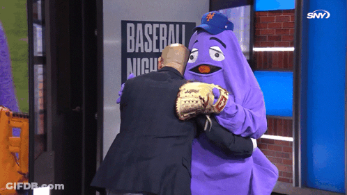 A Man Is Hugging A Purple Mascot In Front Of A Baseball Night Sign GIF