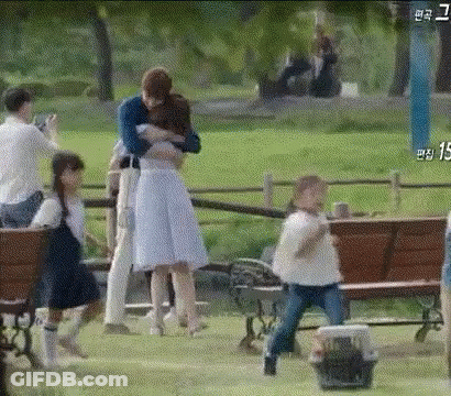 A Man Is Hugging A Woman In A Park While A Group Of People Are Standing Around A Bench . GIF