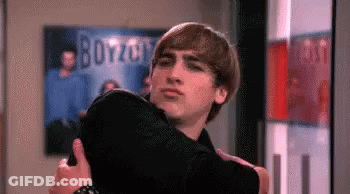 A Man Is Hugging A Woman In A Room With A Poster On The Wall That Says Boyzoo . GIF