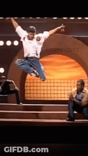 A Man Is Jumping In The Air On A Stage While Another Man Sits On The Steps . GIF
