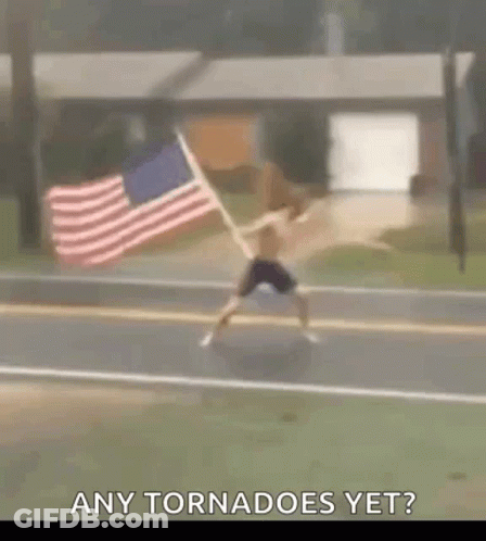 A Man Is Jumping In The Air While Holding An American Flag On The Side Of The Road . GIF