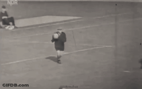 A Man Is Kicking A Soccer Ball On A Field While Another Man Sits On The Ground . GIF