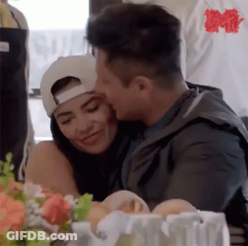 A Man Is Kissing A Woman On The Cheek While Sitting At A Table With Flowers . GIF