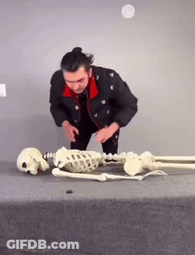 A Man Is Kneeling Down Next To A Skeleton And A Skull On A Table . GIF