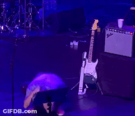 A Man Is Kneeling Down On A Stage With A Guitar Behind Him GIF