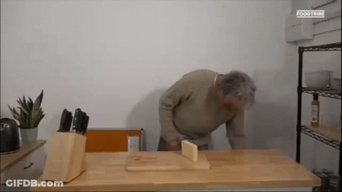 A Man Is Kneeling Down On A Wooden Table With A Knife Block And A Cutting Board . GIF