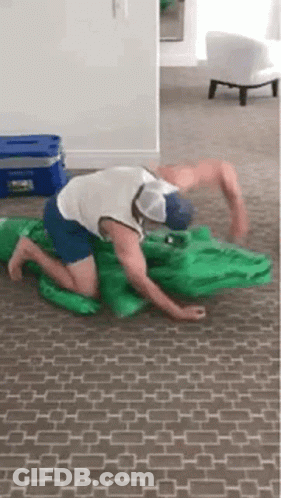 A Man Is Kneeling On The Floor With A Green Inflatable Alligator . GIF