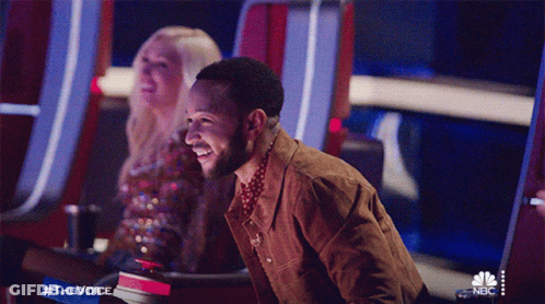 A Man Is Laughing While Sitting In Front Of A Nbc Sign GIF