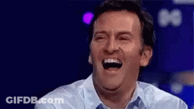 A Man Is Laughing With His Mouth Open While Sitting On A Stage . GIF