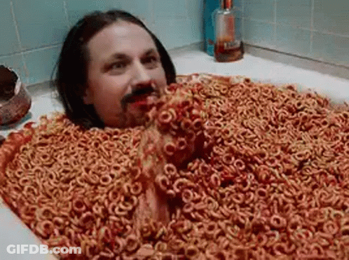 A Man Is Laying In A Bathtub Filled With Cereal Rings . GIF
