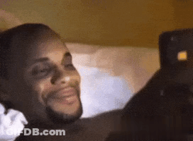 A Man Is Laying On A Bed Looking At A Cell Phone And Smiling . GIF