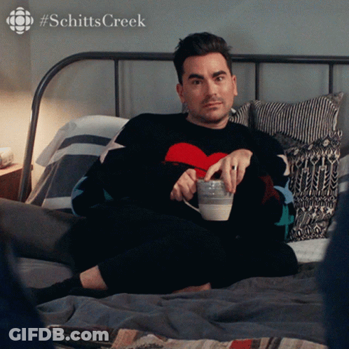 A Man Is Laying On A Bed With A Cup Of Coffee And A #Schitts Creek Logo GIF