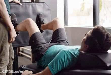 A Man Is Laying On A Bench In A Gym While A Man Stands Behind Him . GIF