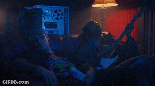 A Man Is Laying On A Couch While Another Man Holds A Guitar GIF