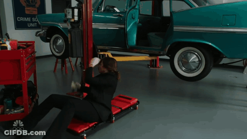 A Man Is Laying On A Rolling Chair In Front Of A Car That Has A Sign That Says Crime Scene Unit On It GIF