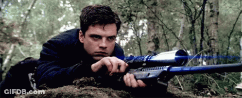 A Man Is Laying On The Ground Holding A Sniper Rifle In A Forest . GIF