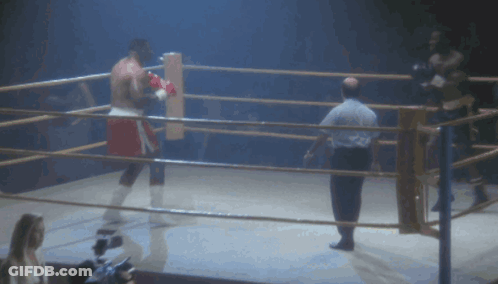 A Man Is Laying On The Ground In A Boxing Ring While Another Man Watches GIF