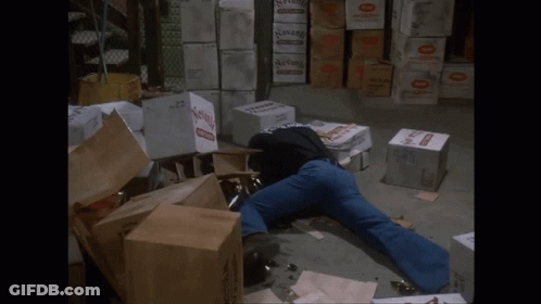A Man Is Laying On The Ground In A Warehouse Surrounded By Boxes Including One That Says Seven GIF