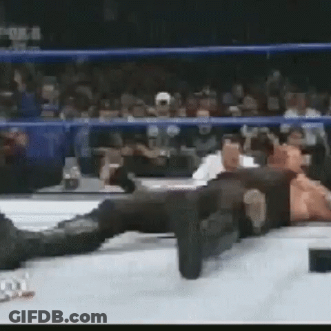 A Man Is Laying On The Ground In A Wrestling Ring While A Crowd Watches . GIF
