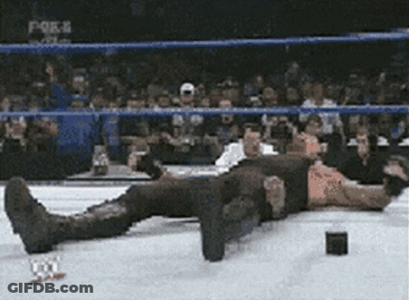 A Man Is Laying On The Ground In A Wrestling Ring While A Crowd Watches . GIF