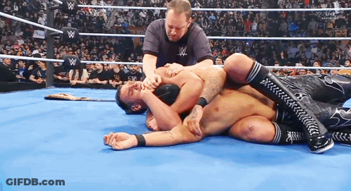 A Man Is Laying On The Ground In A Wrestling Ring While A Referee Watches . GIF