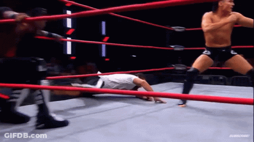 A Man Is Laying On The Ground In A Wrestling Ring While Another Man Is Standing In The Ring . GIF
