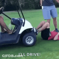 A Man Is Laying On The Ground Next To A Golf Cart While Another Man Stands Behind Him . GIF