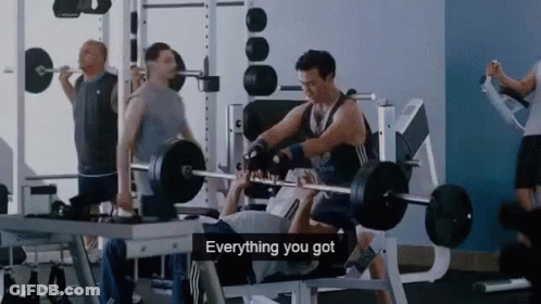 A Man Is Lifting A Barbell In A Gym While Another Man Watches . GIF