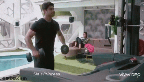 A Man Is Lifting A Dumbbell In A Living Room While A Woman Sits On A Couch . GIF