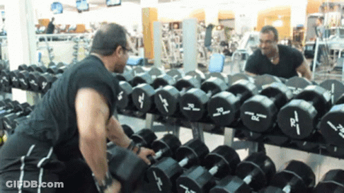 A Man Is Lifting A Dumbbell In Front Of A Row Of Dumbbells With The Letters Dc On Them GIF