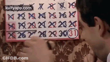 A Man Is Looking At A Calendar On A Wall And Writing On It . GIF