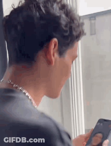 A Man Is Looking At A Cell Phone While Sitting In Front Of A Window . GIF