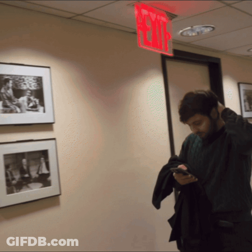 A Man Is Looking At His Phone In A Hallway With A Red Exit Sign Above Him GIF