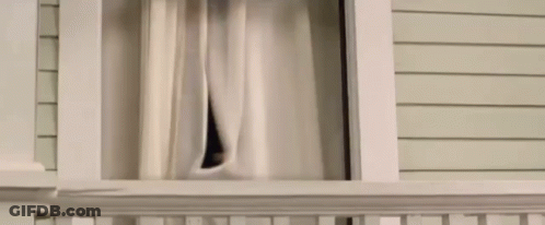 A Man Is Looking Out Of A Window With Curtains . GIF