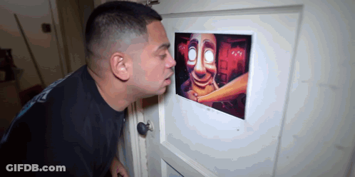 A Man Is Looking Through A Peephole At A Picture Of A Scary Face . GIF