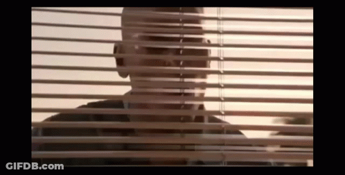 A Man Is Looking Through A Window With Blinds . GIF