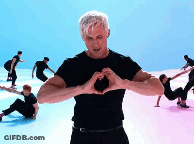 A Man Is Making A Heart Shape With His Hands While A Group Of People Are Dancing In The Background . GIF