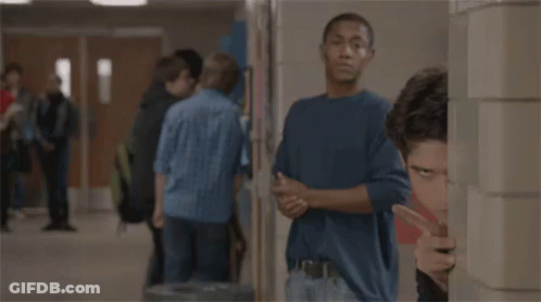 A Man Is Peeking Out From Behind A Wall In A Hallway While A Group Of People Are Standing In The Background . GIF
