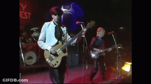 A Man Is Playing A Bass Guitar On A Stage While Another Man Sings Into A Microphone . GIF