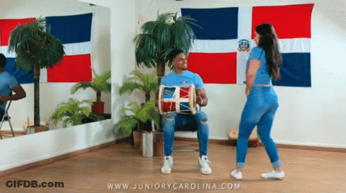 A Man Is Playing A Drum While A Woman Is Dancing In A Room With Flags Behind Them GIF
