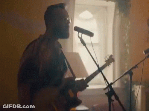 A Man Is Playing A Guitar And Singing Into A Microphone While Standing In Front Of A Window . GIF