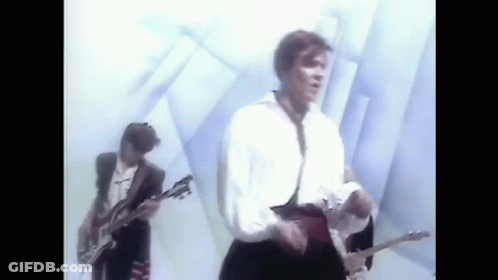 A Man Is Playing A Guitar In A Band While Another Man Sings . GIF