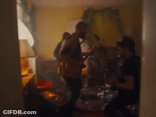 A Man Is Playing A Guitar In A Living Room While Another Man Plays Drums . GIF