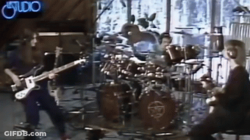 A Man Is Playing A Guitar In Front Of A Drum Set With A Neon Sign That Says Studio GIF